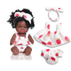 A black-skinned doll with curly hair wearing a white outfit and pink accents, accompanied by a matching bow and diaper cover.