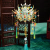 A detailed 3D metal phoenix crown model kit with colorful feathers and jewels displayed in front of a dark wooden screen.