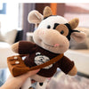 A hand holds a brown and white plush cow stuffed animal with a leather strap.
