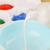 A white bottle of acrylic paint thinner is pouring white liquid into a light blue bowl, with other colored paints visible in 