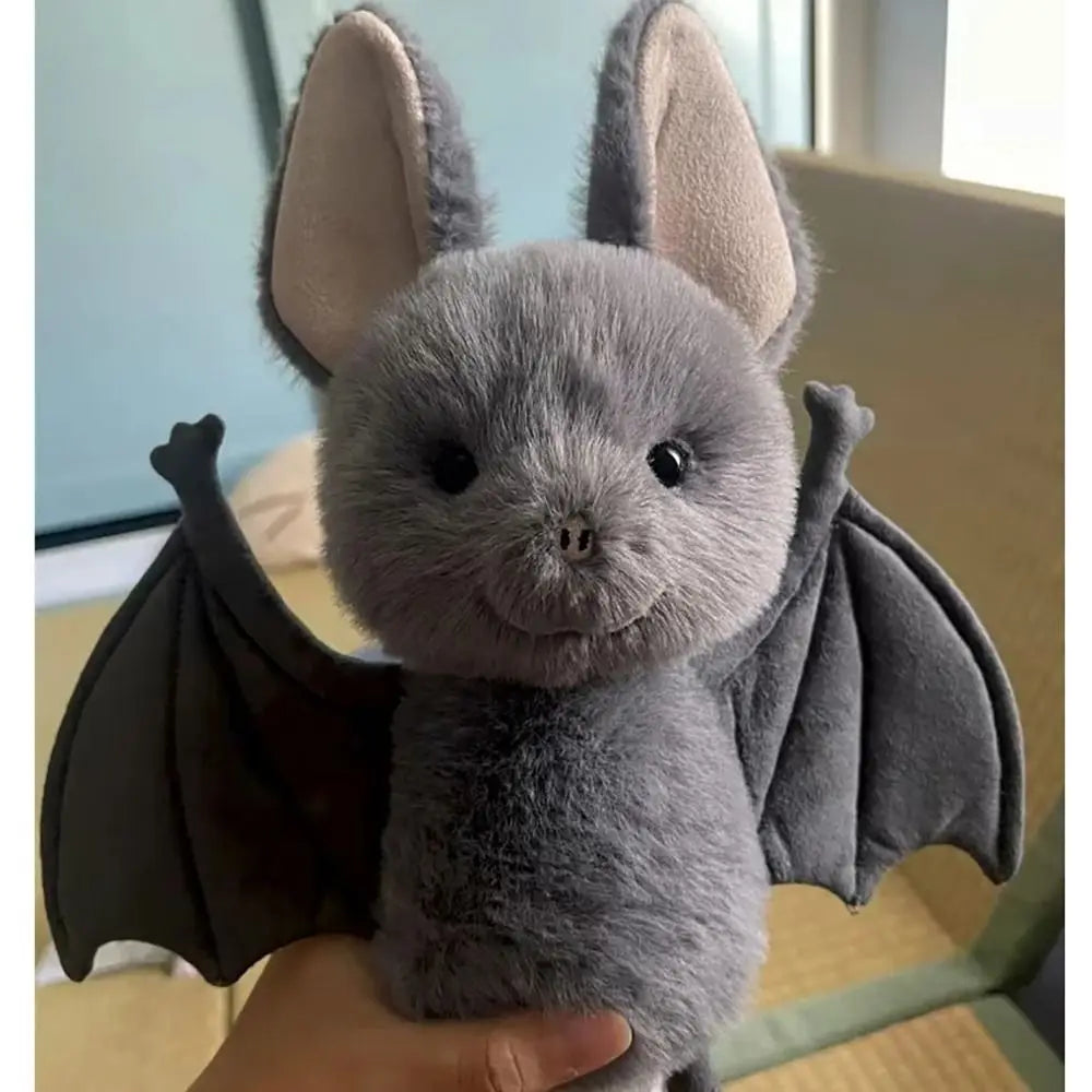 A soft grey bat plush toy with large ears and dark wings is held in a person's hand.