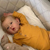 A baby doll with soft cloth body wearing a yellow long-sleeved shirt is lying on white bedding.