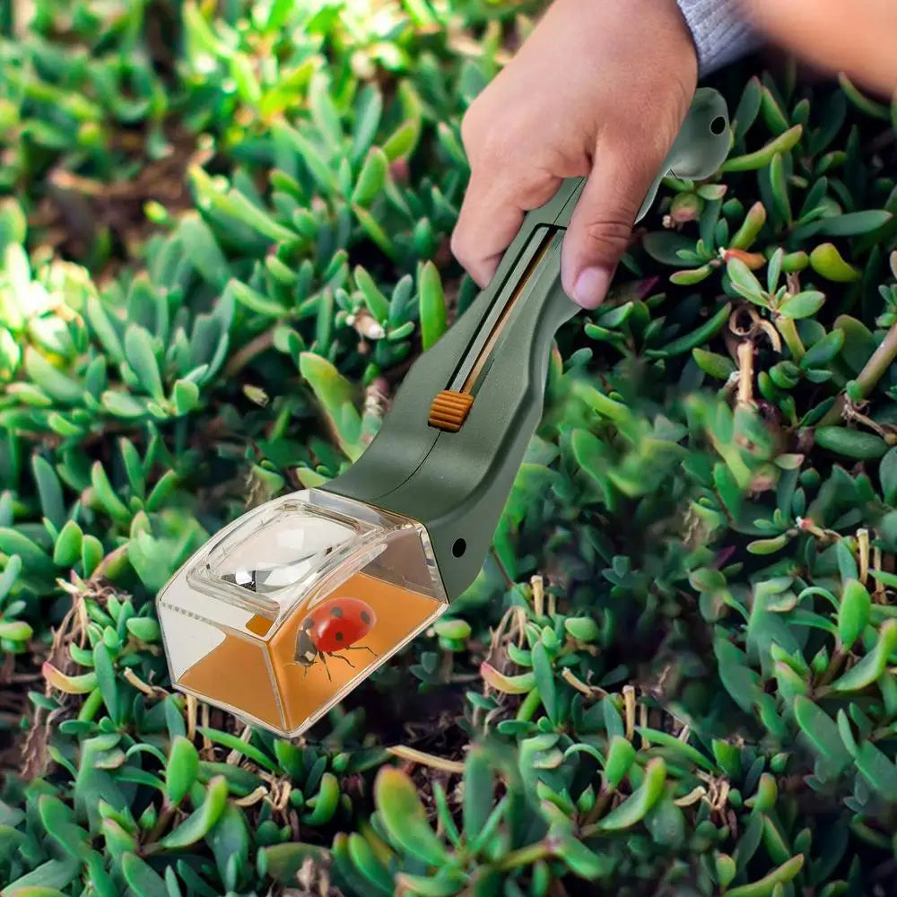 A hand holds a green handheld insect catcher with a magnifying lens in a natural, leafy outdoor setting.