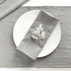 A gray linen dinner napkin set with a sprig of dried foliage placed on a white plate and gray cloth.