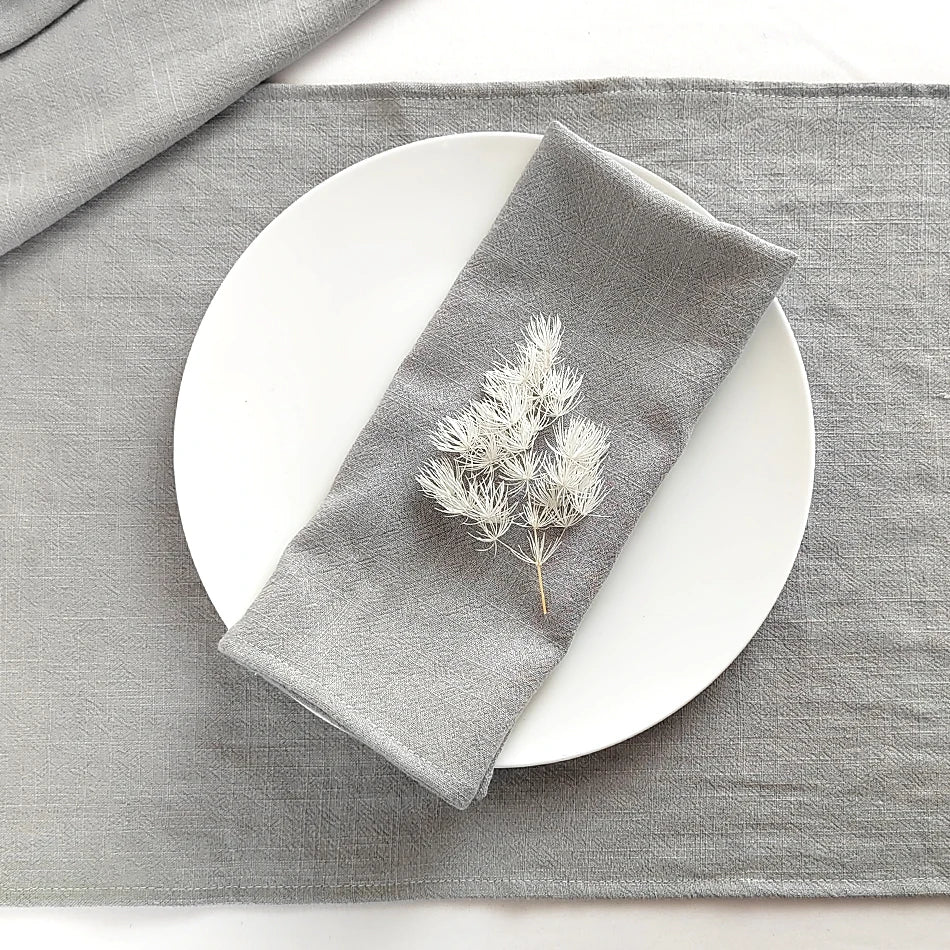 A gray linen dinner napkin set with a sprig of dried foliage placed on a white plate and gray cloth.