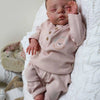 A newborn baby doll with a soft cloth body wearing pink clothing and a light colored bow, resting on a white fluffy surface.