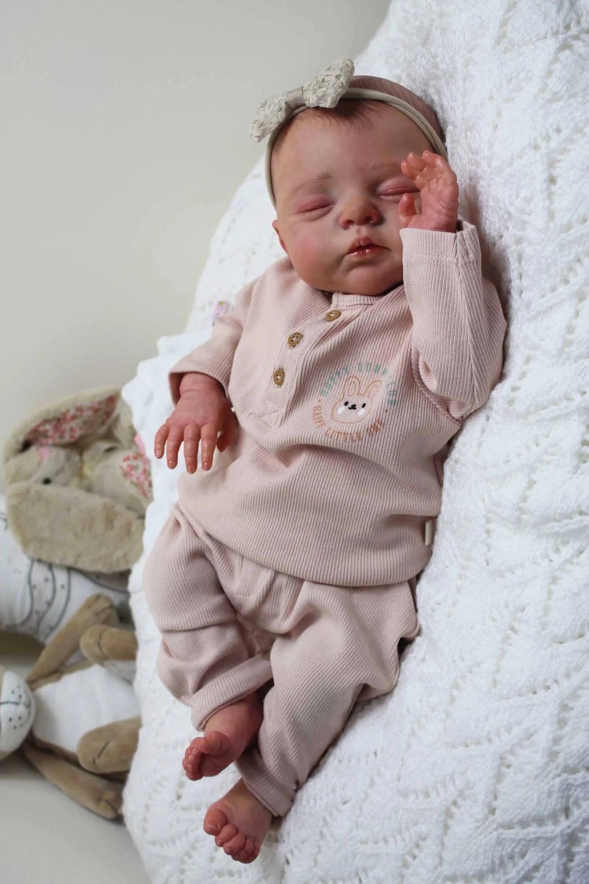 A newborn baby doll with a soft cloth body wearing pink clothing and a light colored bow, resting on a white fluffy surface.