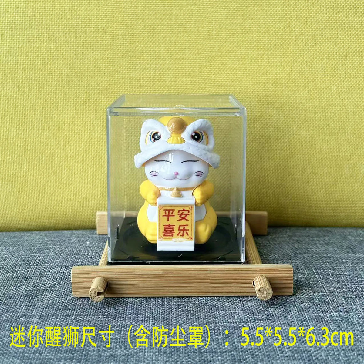 A yellow cat figurine with a white hat is displayed in a clear glass box on a wooden base.