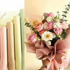 A bouquet of pink and cream roses and greenery wrapped in soft blush paper, next to several rolls of translucent tissue paper