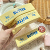 A hand holds a yellow butter stick fidget toy with the label Butter and 4 oz net weight.