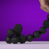 A hand is holding a string of black soft sensory silicone massage balls with magnetic cores against a purple background.