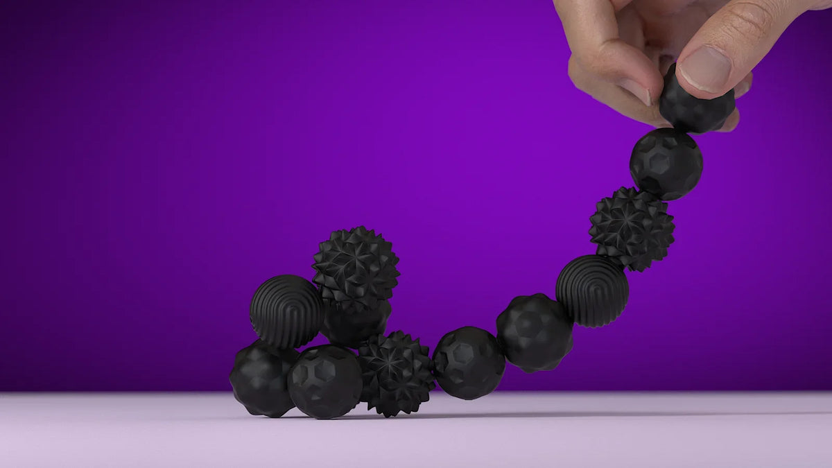 A hand is holding a string of black soft sensory silicone massage balls with magnetic cores against a purple background.
