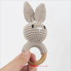 A handmade knitted bunny rattle and teether ring made of light gray yarn, held by a hand.