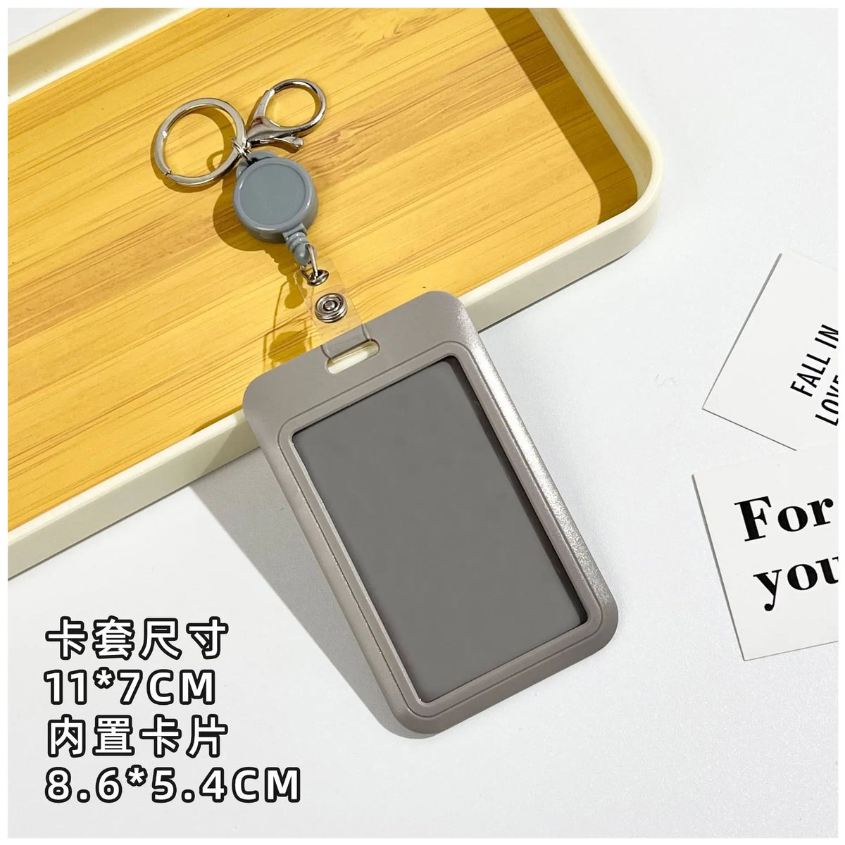A gray retractable ID badge holder with a clip is shown on a wooden tray next to a keychain.