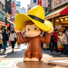 A collectible figure of Curious George wearing a yellow cowboy hat, standing on a wooden surface.