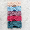 A stack of five pastel colored fabric bow baby headbands in shades of peach, blue, mauve, pink, and teal.