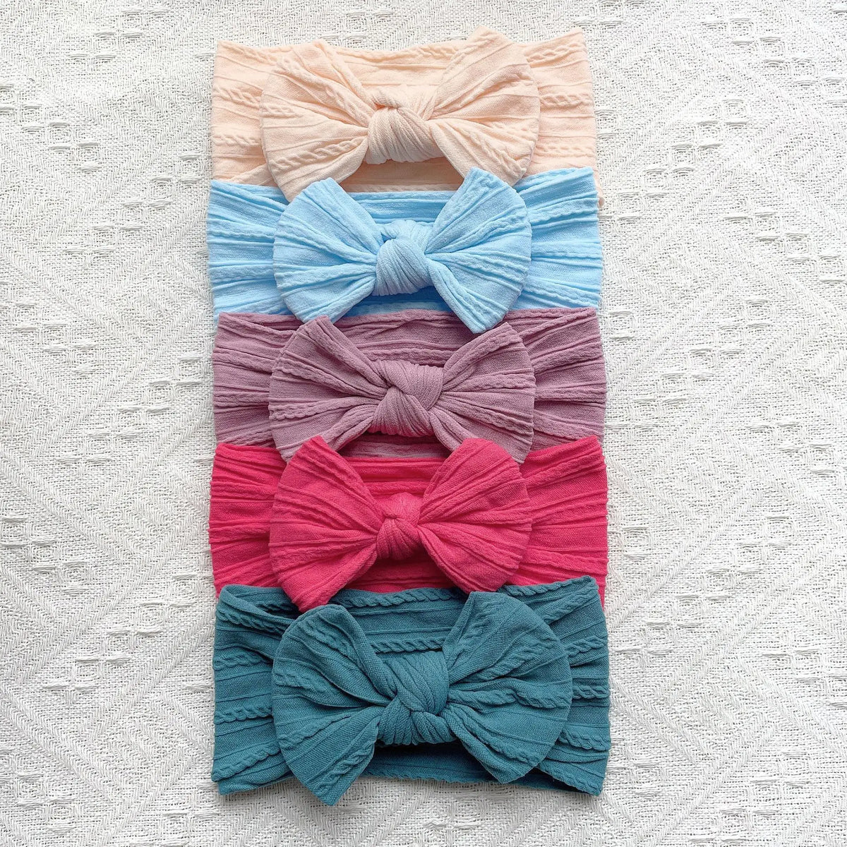 A stack of five pastel colored fabric bow baby headbands in shades of peach, blue, mauve, pink, and teal.