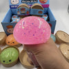 A hand holds a large, pink, glittery squishy dumpling toy next to other similar toys in their packaging.