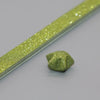 A close-up of a thin strip of glitter origami star paper, sparkling gold green, lying on a gray surface.