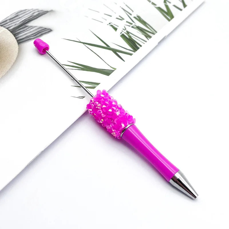 A bright pink sequin-covered ballpoint pen is shown resting on a white surface next to green foliage.