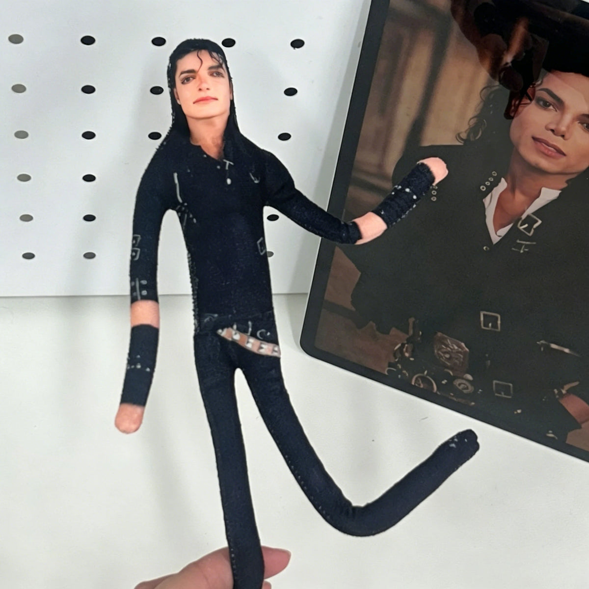 A dark clothing doll dressed in a black jumpsuit and boots, posed next to a photo of Michael Jackson.