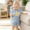 A baby boy wearing a blue short-sleeve romper with yellow bear and mushroom appliqués sits on a sofa.