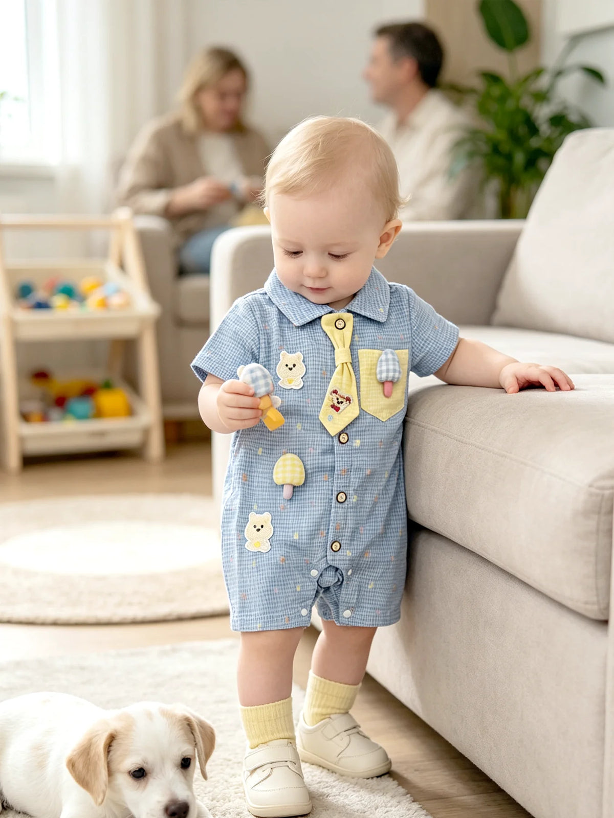 A baby boy wearing a blue short-sleeve romper with yellow bear and mushroom appliqués sits on a sofa.