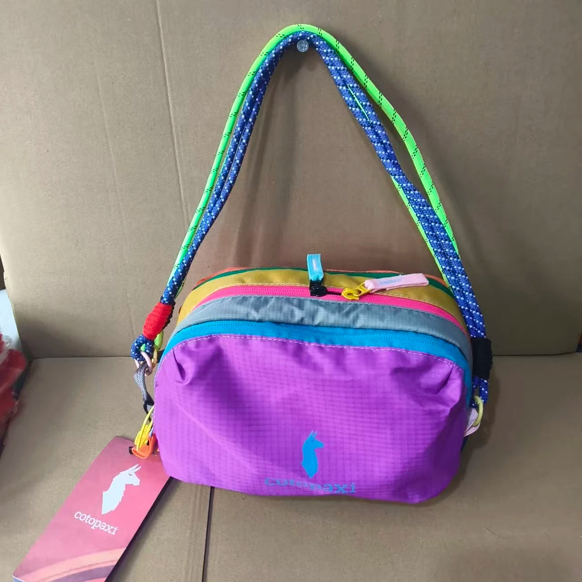 A purple alpaca themed travel storage bag with a multicolored strap is shown against a brown cardboard background.