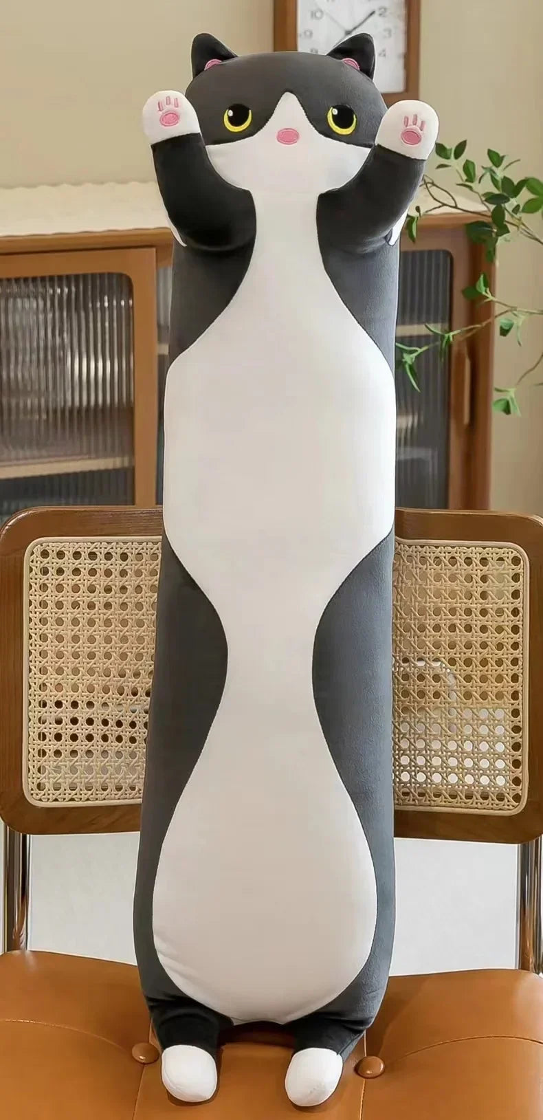 A plush cat pillow resembling a black and white cat is sitting upright on a brown leather sofa.