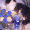 A black and white cat is resting on a white surface next to a glass of blue liquid and flowers.
