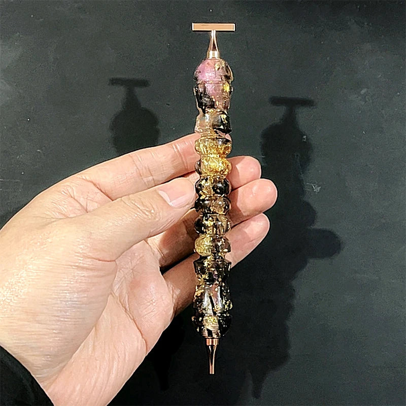 A hand holds a colorful diamond painting pen with a stainless steel tip, displaying various iridescent and dark colored stone