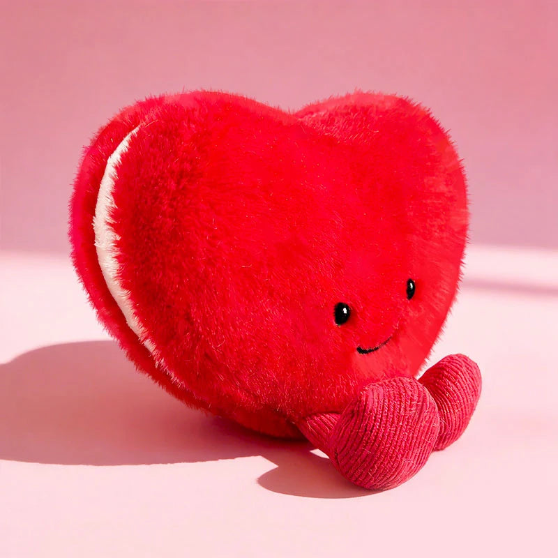 A bright red heart-shaped plush cushion with a white trim, smiling expression, resting on a pink surface.