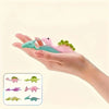 A hand holds several colorful mini dinosaur finger slingshots, including pink, green, and purple varieties.