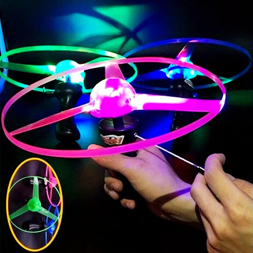 A hand holds a glowing pink UFO spinning top with a pull string, demonstrating the LED flying disc toy.