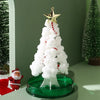 A white flocked Christmas tree decoration topped with a gold star, placed on a green circular base next to a Santa figurine.