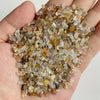A hand holds a pile of natural tumbled stone chips in various shades of amber, brown, and clear quartz.
