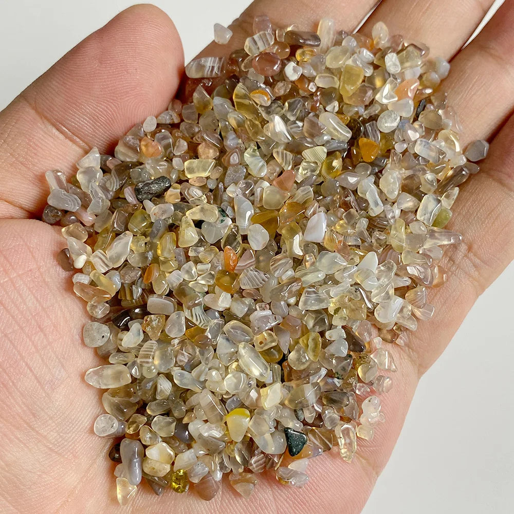 A hand holds a pile of natural tumbled stone chips in various shades of amber, brown, and clear quartz.