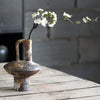 A vintage handcrafted ceramic vase holds a sprig of white flowers on a weathered wooden surface.