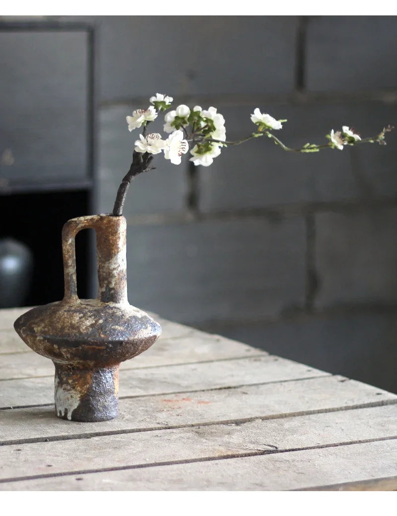 A vintage handcrafted ceramic vase holds a sprig of white flowers on a weathered wooden surface.