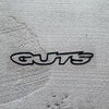 A black vinyl sticker with the word GUTS is placed on a textured gray surface.