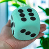 A hand holds a light mint green jumbo soft foam die with black dots, set against a blurred indoor background.
