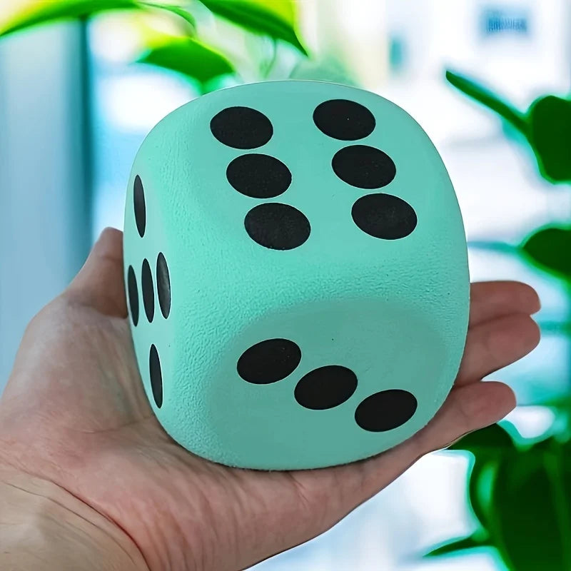 A hand holds a light mint green jumbo soft foam die with black dots, set against a blurred indoor background.