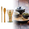 A set of traditional bamboo matcha whisks and a dark ceramic tea bowl with a lid on a wooden tray.
