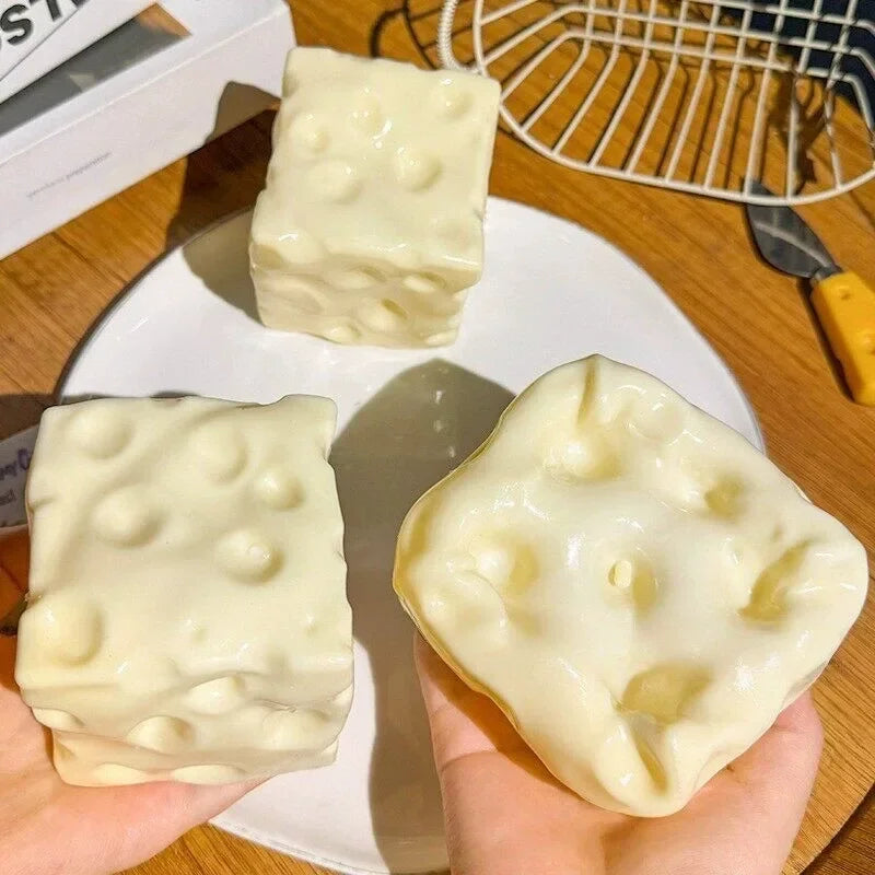 A hand holds a white cheese cube stress ball, a deformable sensory fidget toy, on a white plate.