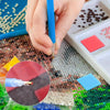 A hand applies a blue tool to diamond painting clay on a mosaic surface next to a white tray of colored stones.