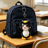 A black backpack with a cute penguin plush toy hanging from the zipper pull, resting on a wooden desk.