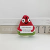 A handwoven crochet ornament shaped like a red watermelon with white eyes and a black mouth, displayed on a light wooden surf