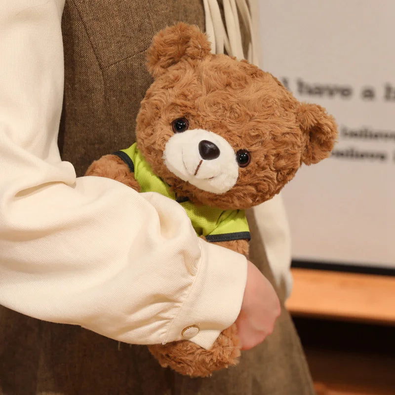 A person is holding a brown teddy bear plush toy with a green shirt in a light-colored sleeve.