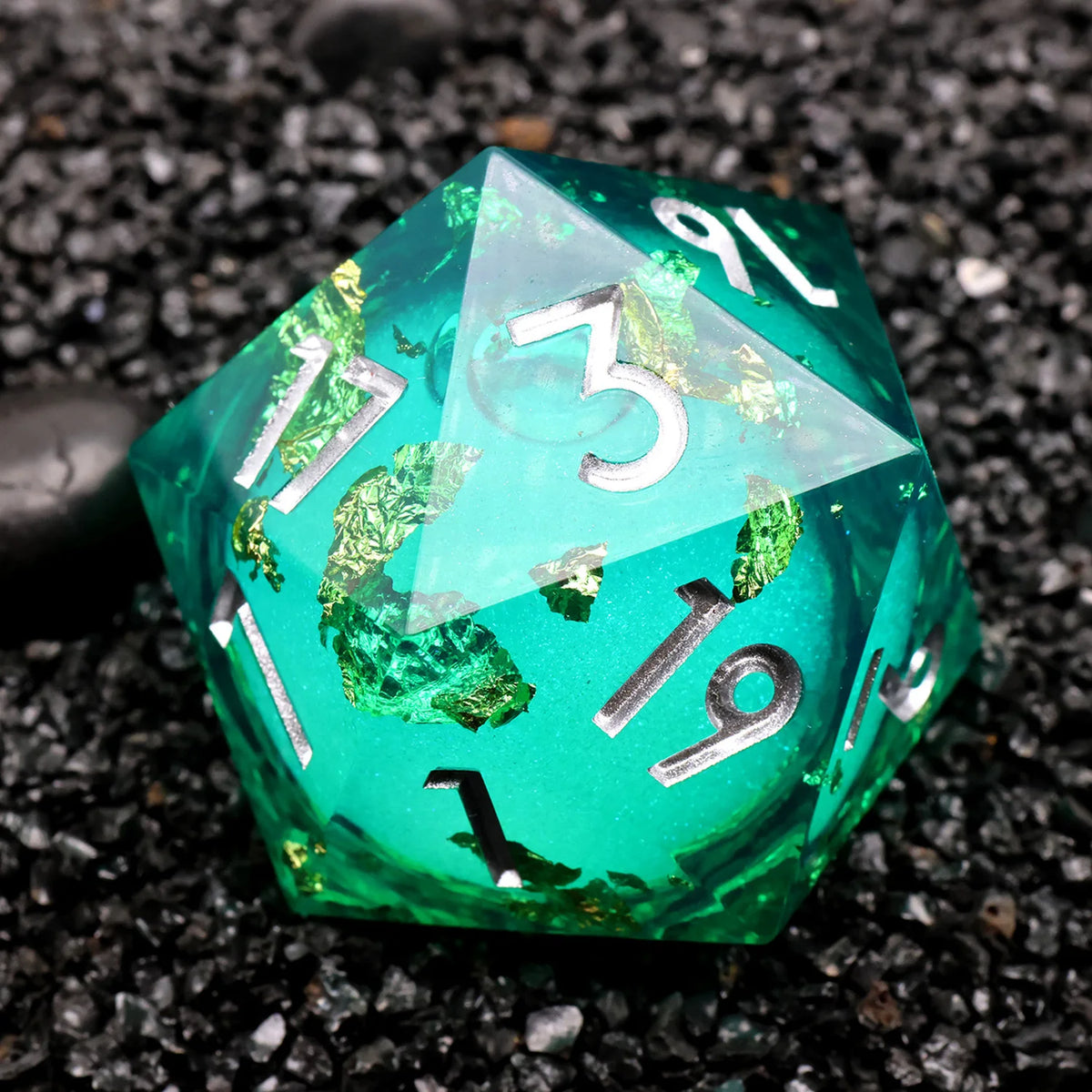 A vibrant green resin D20 die with silver numbers and iridescent liquid swirls resting on black gravel.