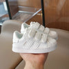 A hand holds a pair of white Adidas Superstar sneakers with hook-and-loop closure.